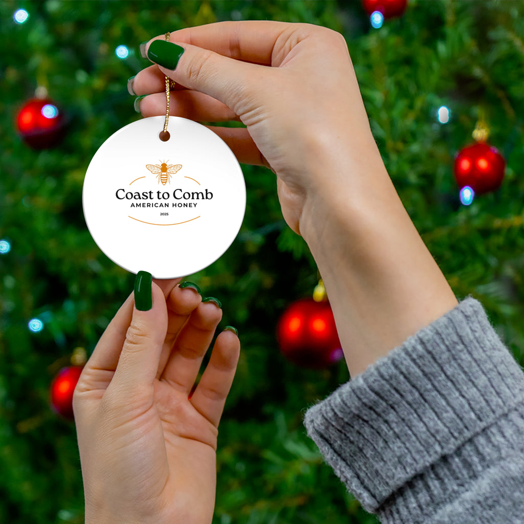 Hand holding a 'Coast to Comb American Honey' ornament against a Christmas tree background.