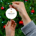 Hand holding a 'Coast to Comb American Honey' ornament against a Christmas tree background.